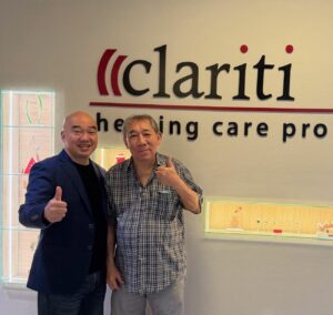 Two men pose indoors, giving thumbs up in front of a wall featuring the red-and-black 'clariti' logo and the slogan 'healing care professionals'.