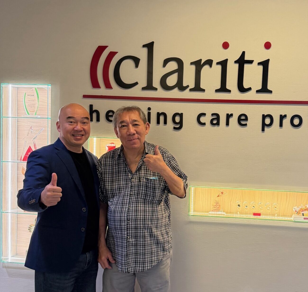 Two men pose indoors, giving thumbs up in front of a wall featuring the red-and-black 'clariti' logo and the slogan 'healing care professionals'.