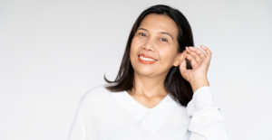 Smiling woman in a white blouse tilting her head slightly and cupping her ear as if listening.