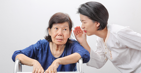 Caregiver whispering to an elderly woman seated in a wheelchair in a bright room.